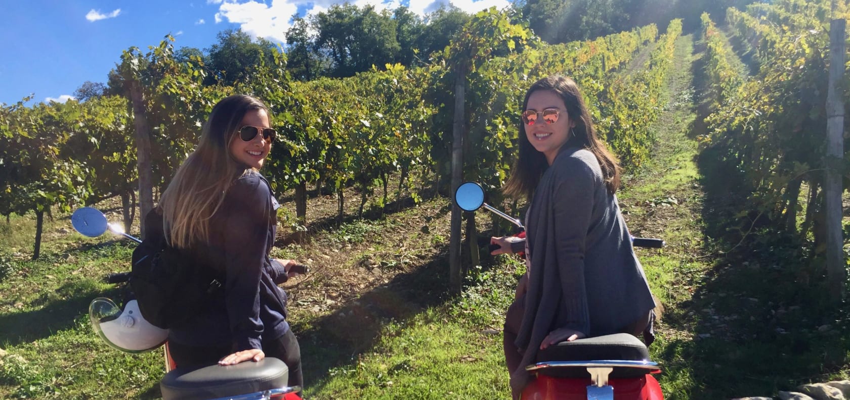 Two girls smiling on mopeds.